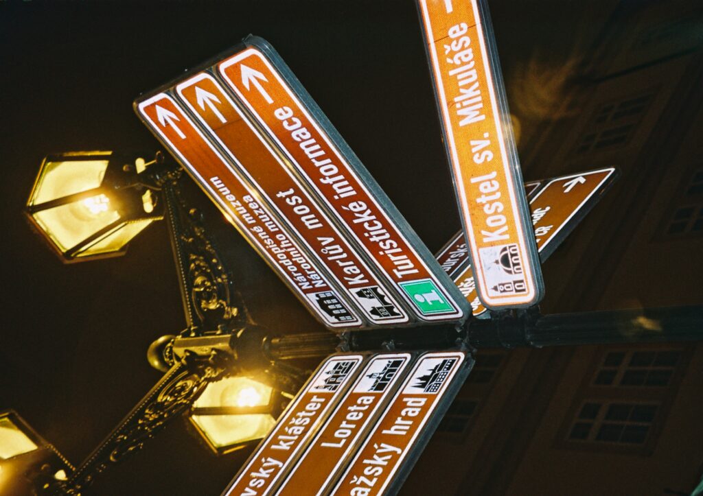 In questa foto è presente un lampione al quale sono appesi vari cartelli con varie indicazioni stradali per i punti di interesse della città.