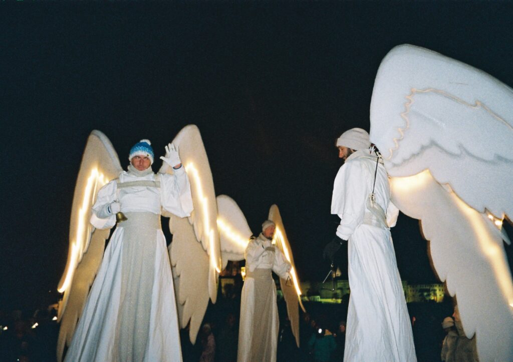 Questa foto rappresenta tre uomini vestiti da angeli con tanto di  ali luminose e trampoli che camminano sul ponte Carlo di notte.