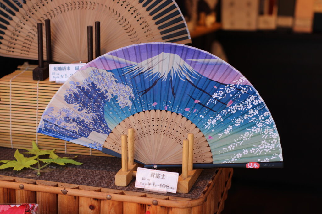 La foto rappresenta un ventaglio giapponese di legno con un decoro molto colorato che rappresenta il monte fuji, l'onda di Hokusai e dei ciliegi in fiore.